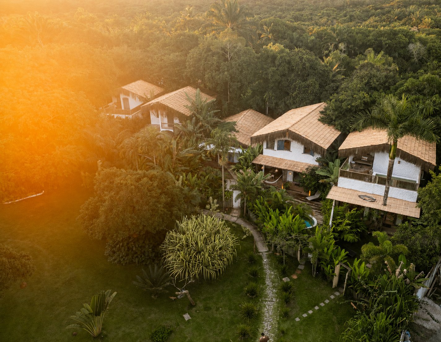 Vista aérea do Bahia Bonita Casas no fim de tarde — telhados de palha e cerâmica entre coqueiros e mata atlântica, jardim ajardinado com caminho de pedras, luz dourada do pôr do sol