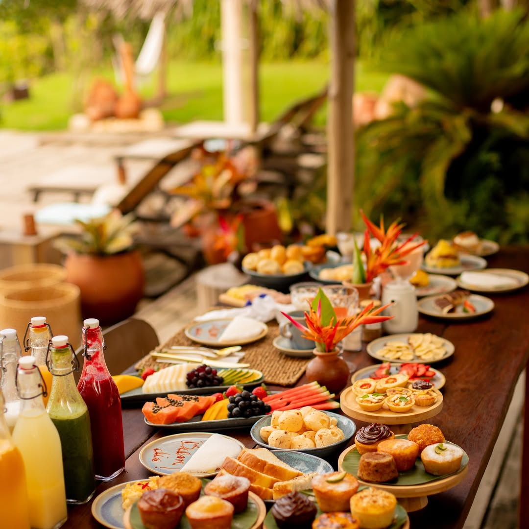 Mesa farta do café da manhã do Bahia Bonita, sob o jardim, sucos prensados em garrafas, frutas, pães, pão de queijo, doces