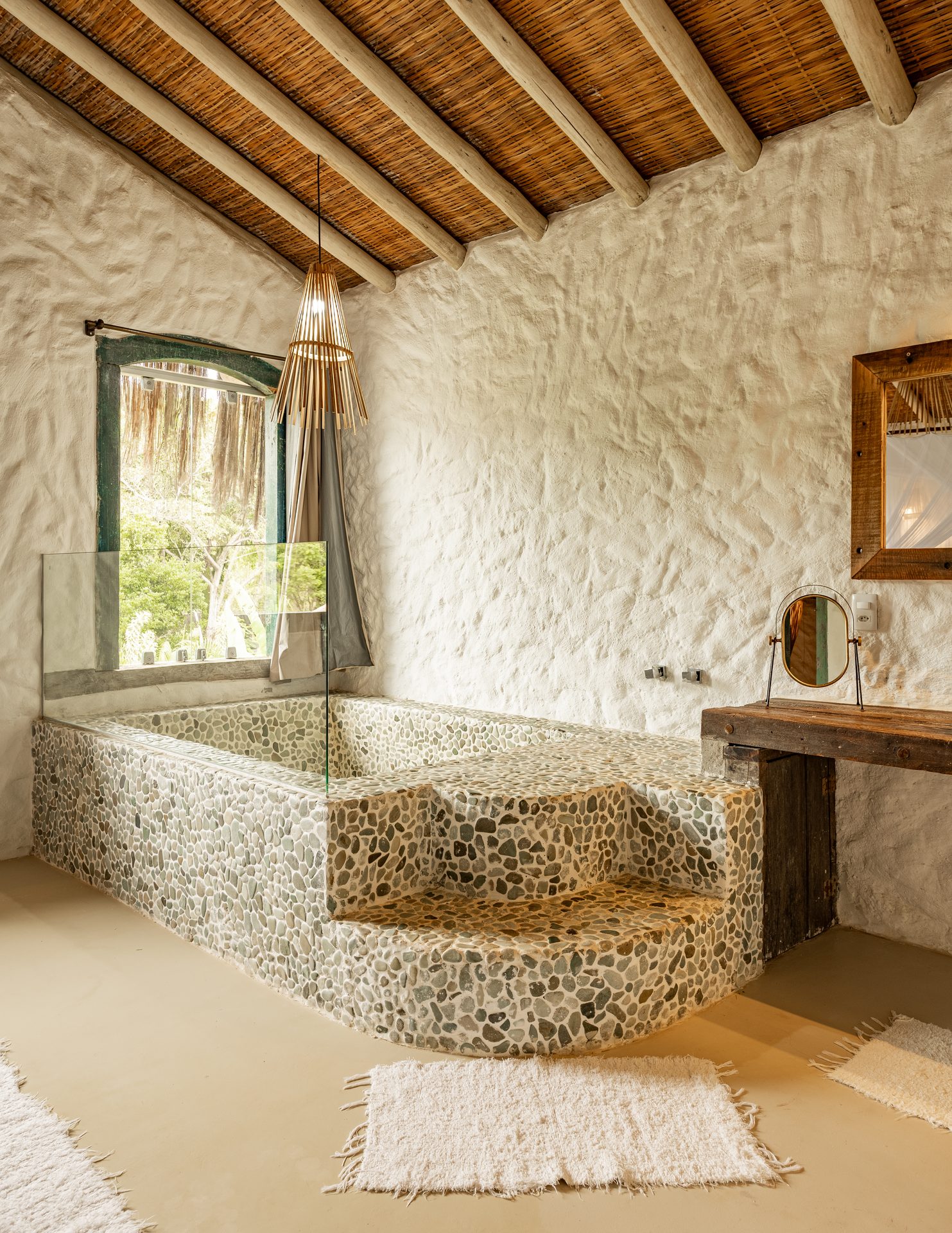 Banheiro da Casa Tupã, banheira revestida com pedrinhas, parede em reboco branco, teto de bambu e madeira