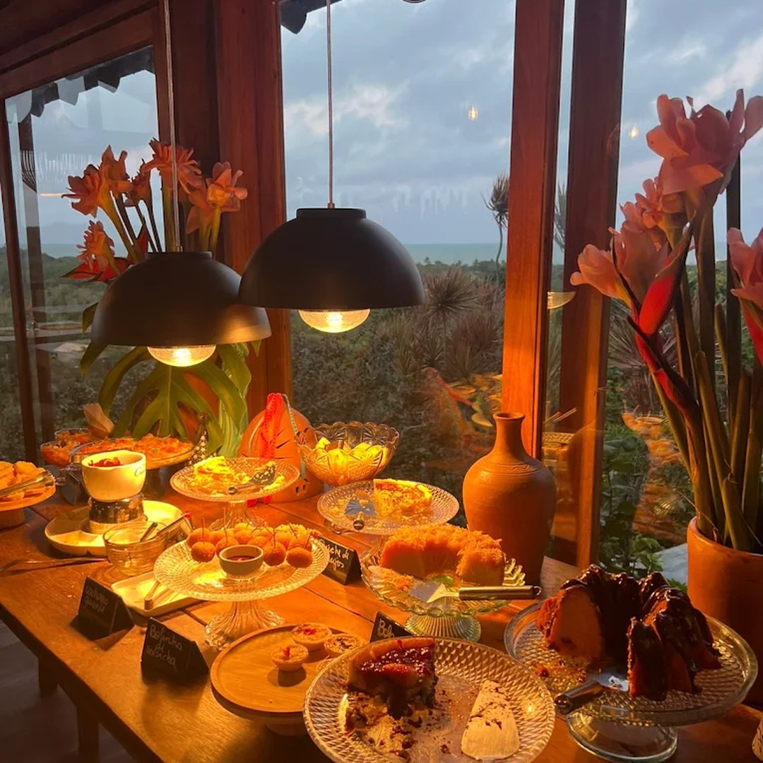 Mesa do chá da tarde com bolos, salgados e geleias caseiras, ao fundo a vista da mata pela janela na luz dourada do fim de tarde