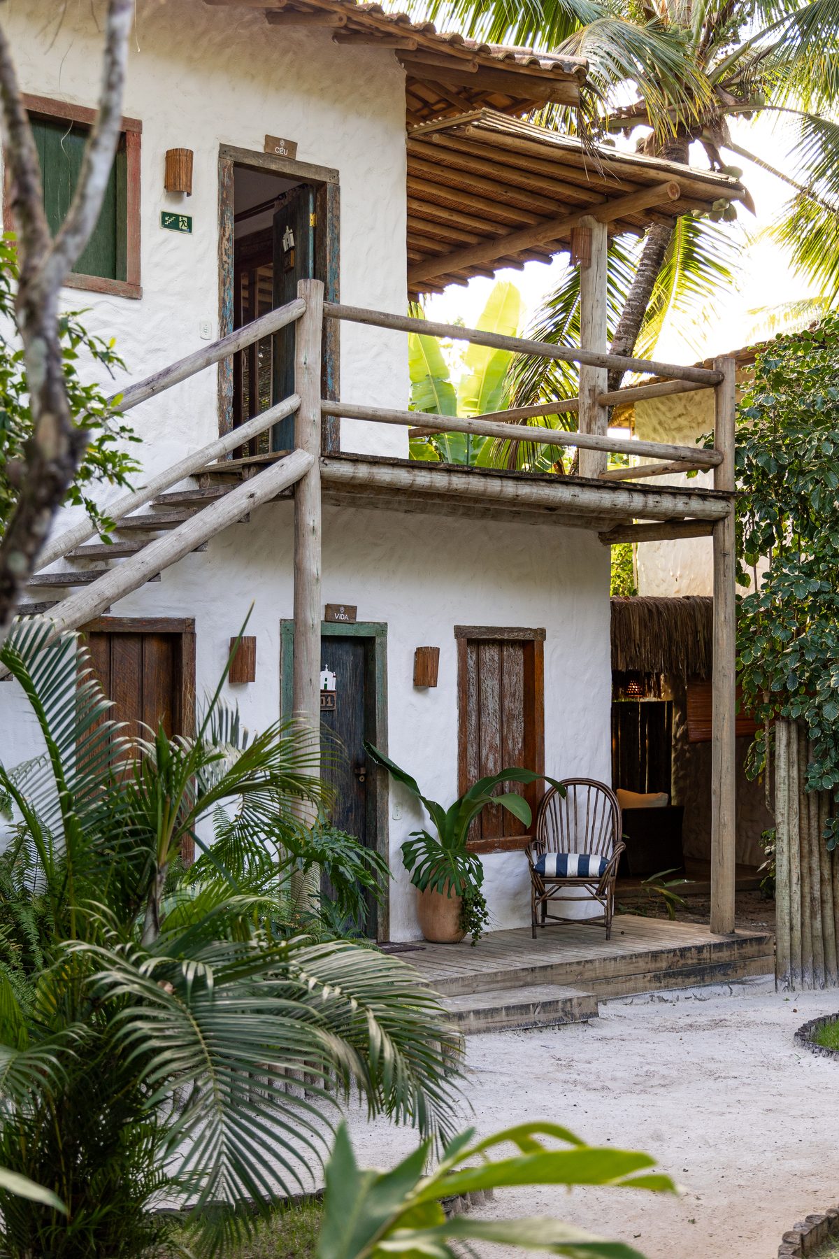 Fachada das suítes do Hotel Boutique Bahia Bonita — arquitetura baiana contemporânea, paredes brancas, portas de madeira de demolição com nomes (Céu, Vida), varanda superior com guarda-corpo rústico e vegetação tropical em volta