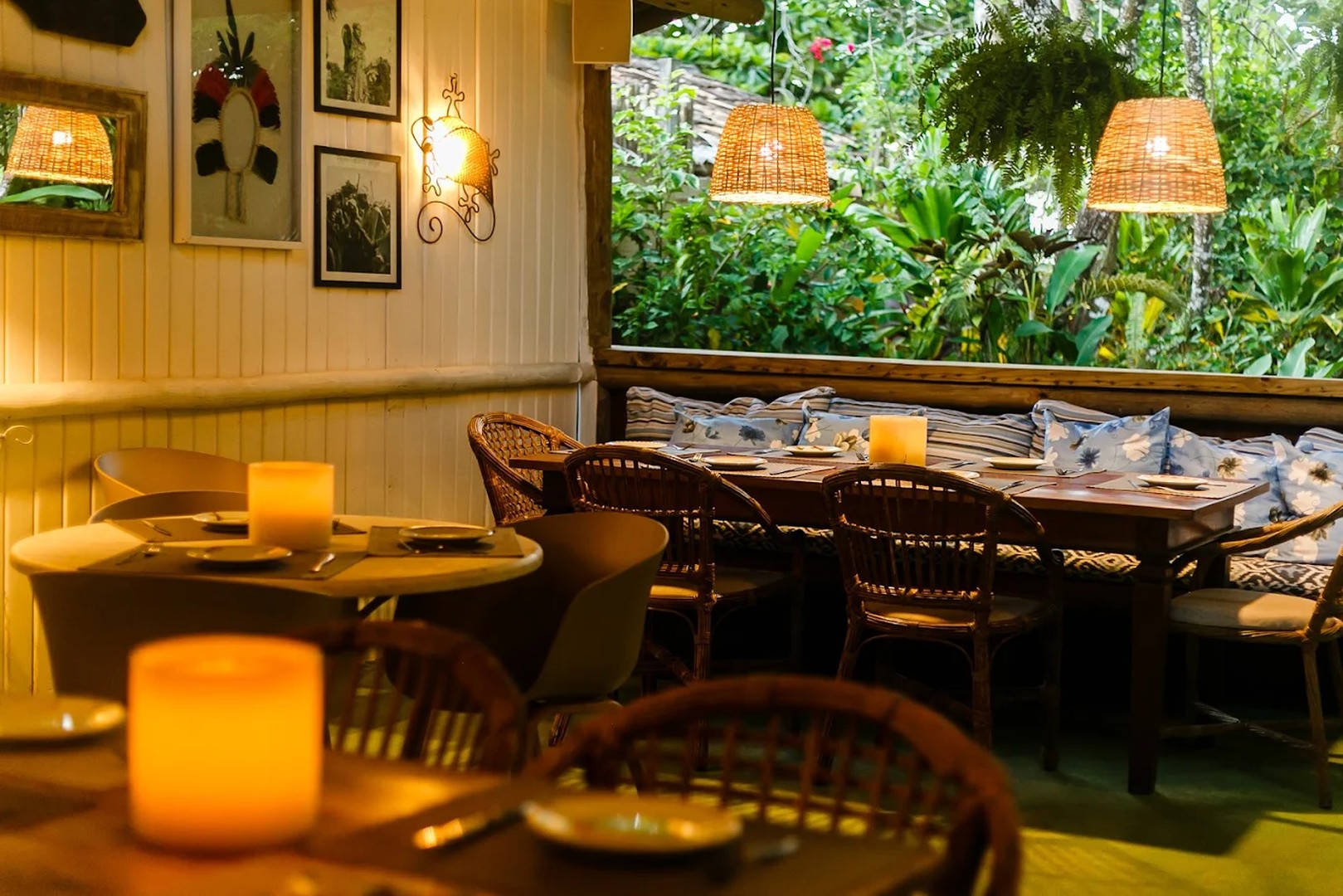 Salão de restaurante em Trancoso à noite, mesas com velas acesas, cadeiras de palhinha e bancos com almofadas estampadas, mata atlântica ao fundo pela janela