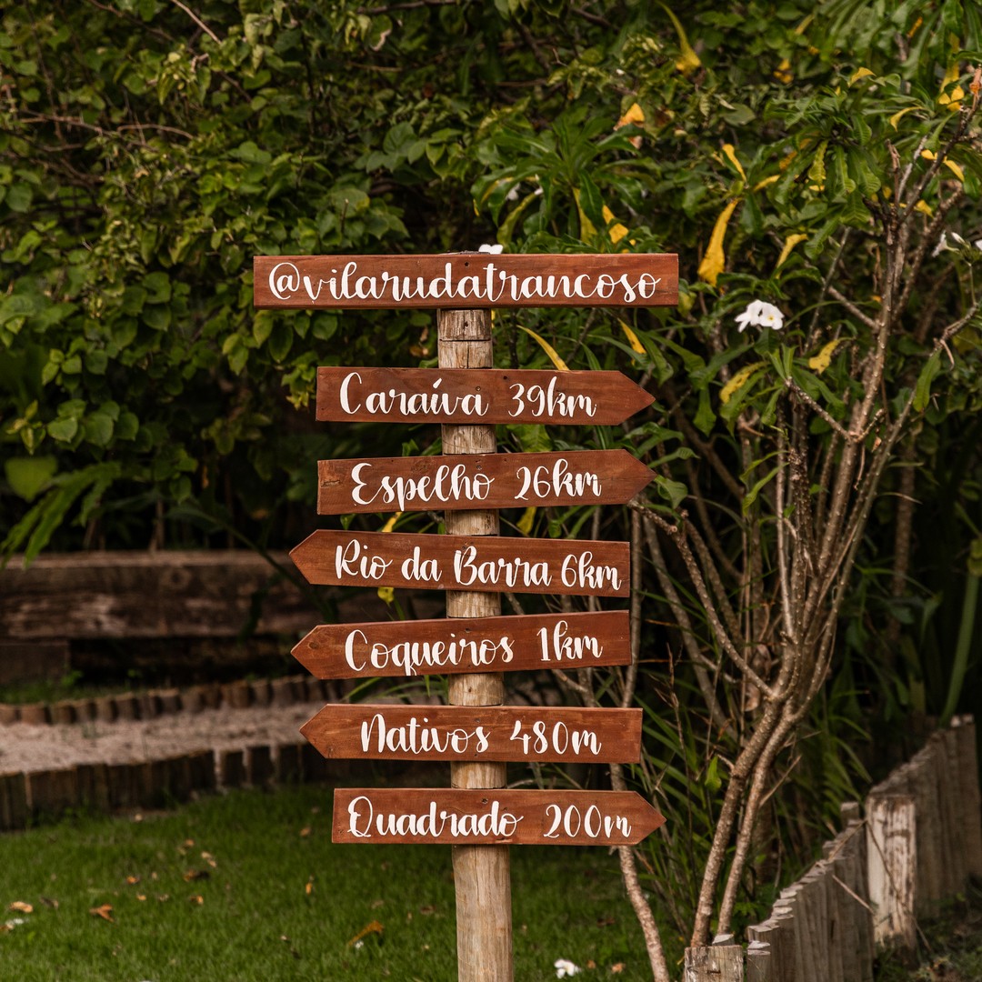 Placa de madeira no jardim do hotel apontando distâncias para praias e pontos de Trancoso — Quadrado 200m, Nativos 480m, Coqueiros 1km, Espelho 26km