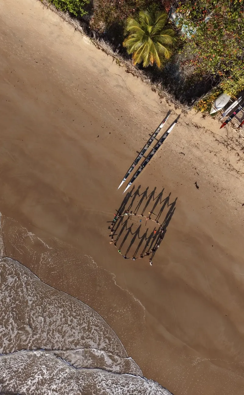Vista aérea do grupo em roda na areia ao lado das canoas